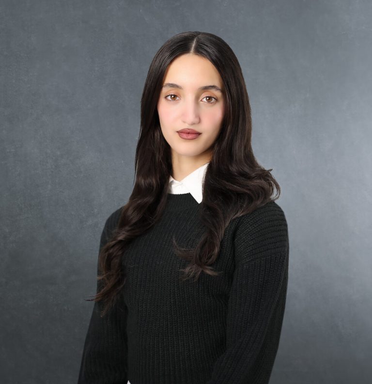 A dark professional photography studio background and a smiling woman wearing a black sweater and a white shirt looking into the camera. Profile photo of Iqra Pasha, legal assistant at Axis Solicitors.
